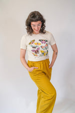 Person wearing a t-shirt with butterfly design and mustard yellow pants on a plain background