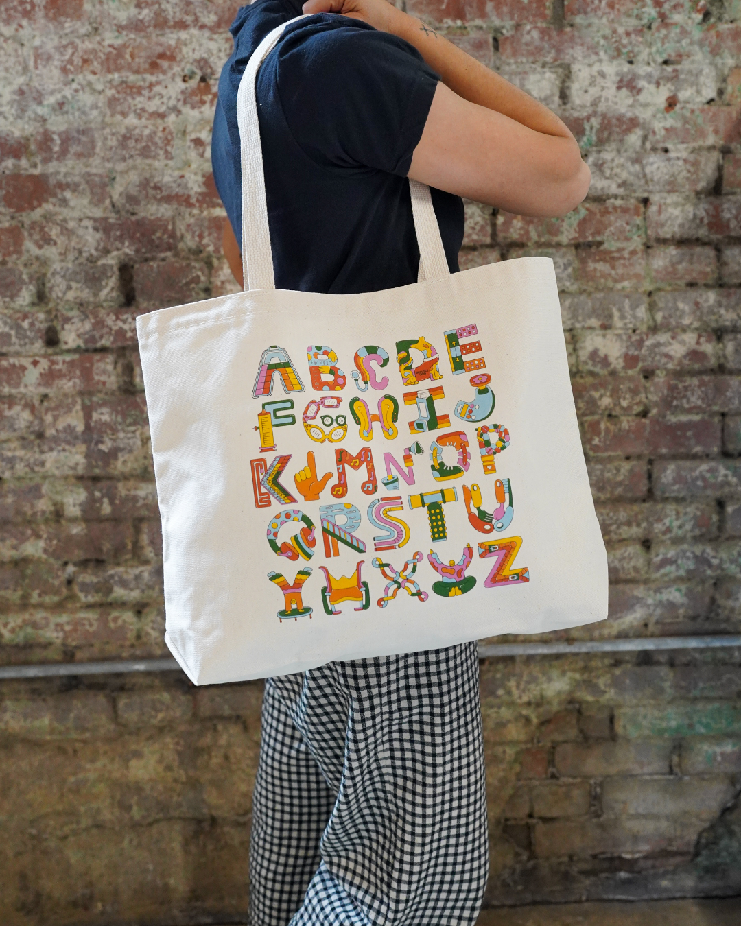 Person holding a tote bag with colorful letters against a brick wall.