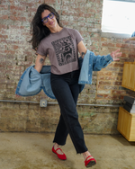 Woman in a casual setting wearing a graphic t-shirt, black pants, red shoes, and a denim jacket over her shoulder.