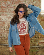 Woman wearing a denim jacket, white shirt with red design saying "We Will Take Care of Each Other", and red pants against a brick wall.