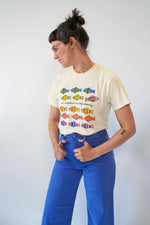 Person wearing a cream t-shirt with colorful fish graphic and blue pants against a white background