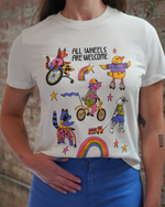 Person wearing a white t-shirt with colorful illustrations and text 'All Wheels Are Welcome' against a brick wall.