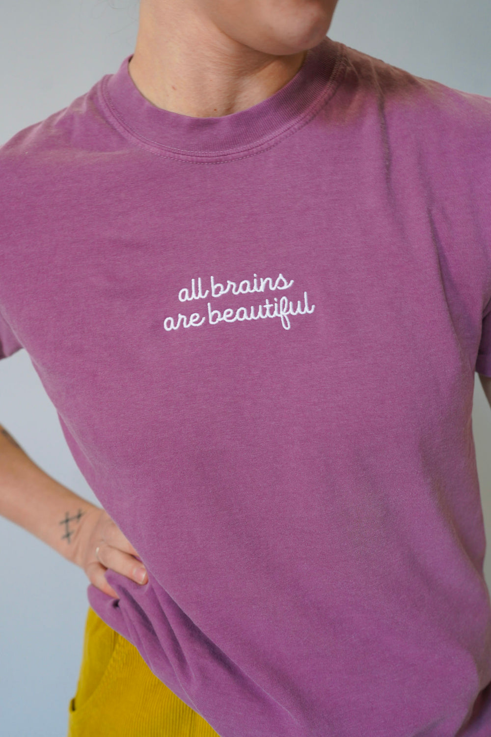 Person wearing a purple t-shirt with 'all brains are beautiful' text on a plain background
