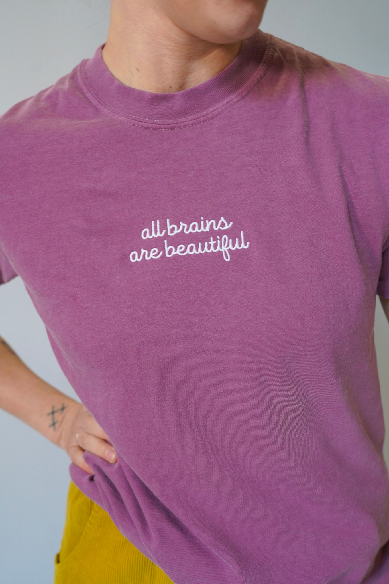 Person wearing a purple t-shirt with 'all brains are beautiful' text on a plain background