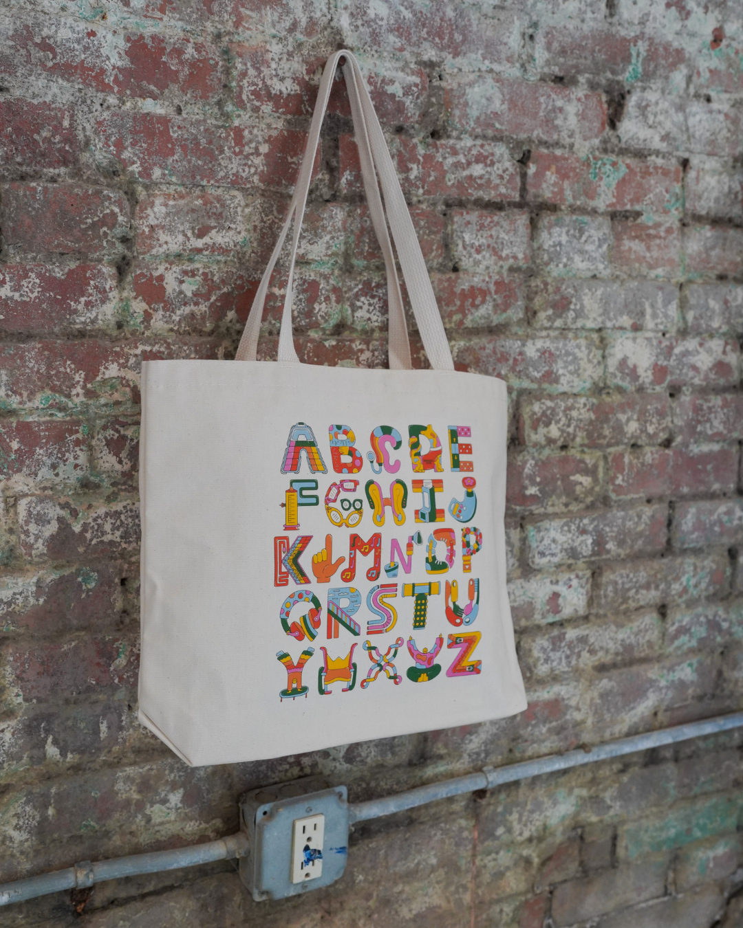 Tote bag with colorful alphabet letters hanging on a brick wall.