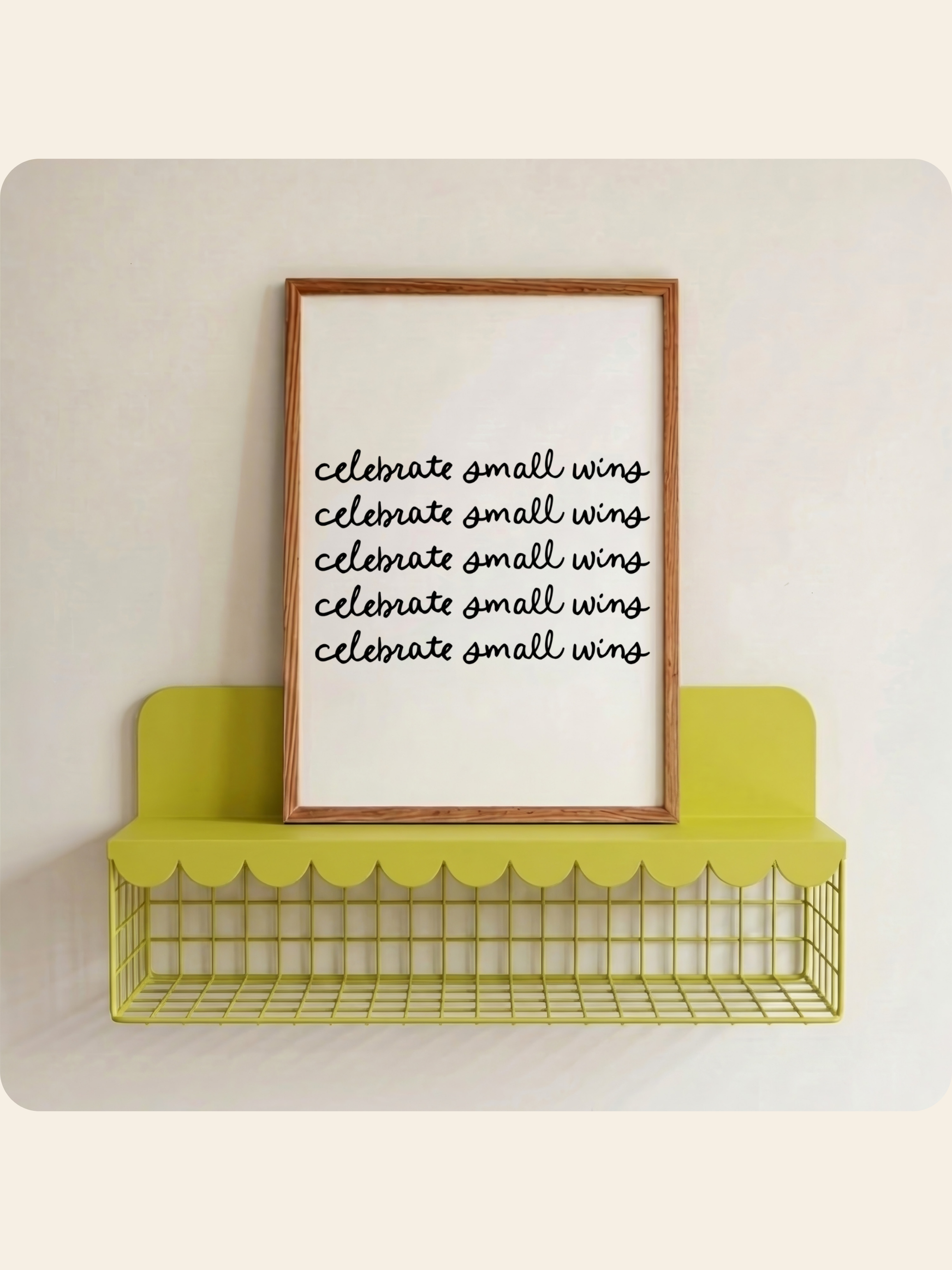 A framed wall sign with the phrase 'celebrate small wings' repeated in black script on a white background, leaning on a yellow wire shelf against a light wall.
