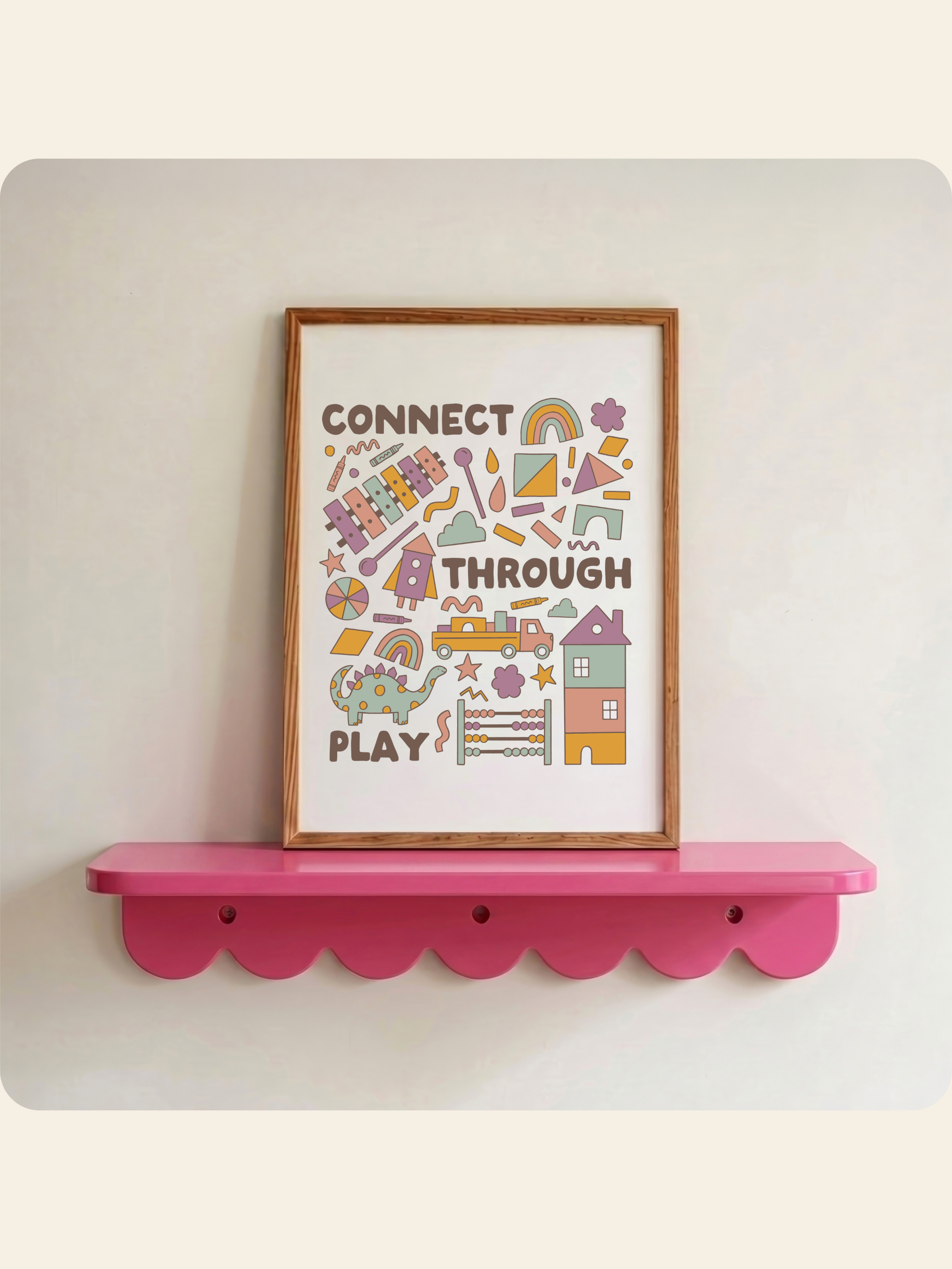 A framed art print with the words 'CONNECT THROUGH PLAY' and colorful playful illustrations, displayed on a bright pink scalloped shelf against a light beige wall.