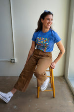 A woman sitting on a wooden stool, wearing a blue t-shirt with colorful text and light brown pants. She has long hair and is smiling, with a neutral background and soft lighting.