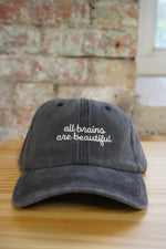 Gray cap with 'all brains are beautiful' text on a wooden surface with a textured wall background