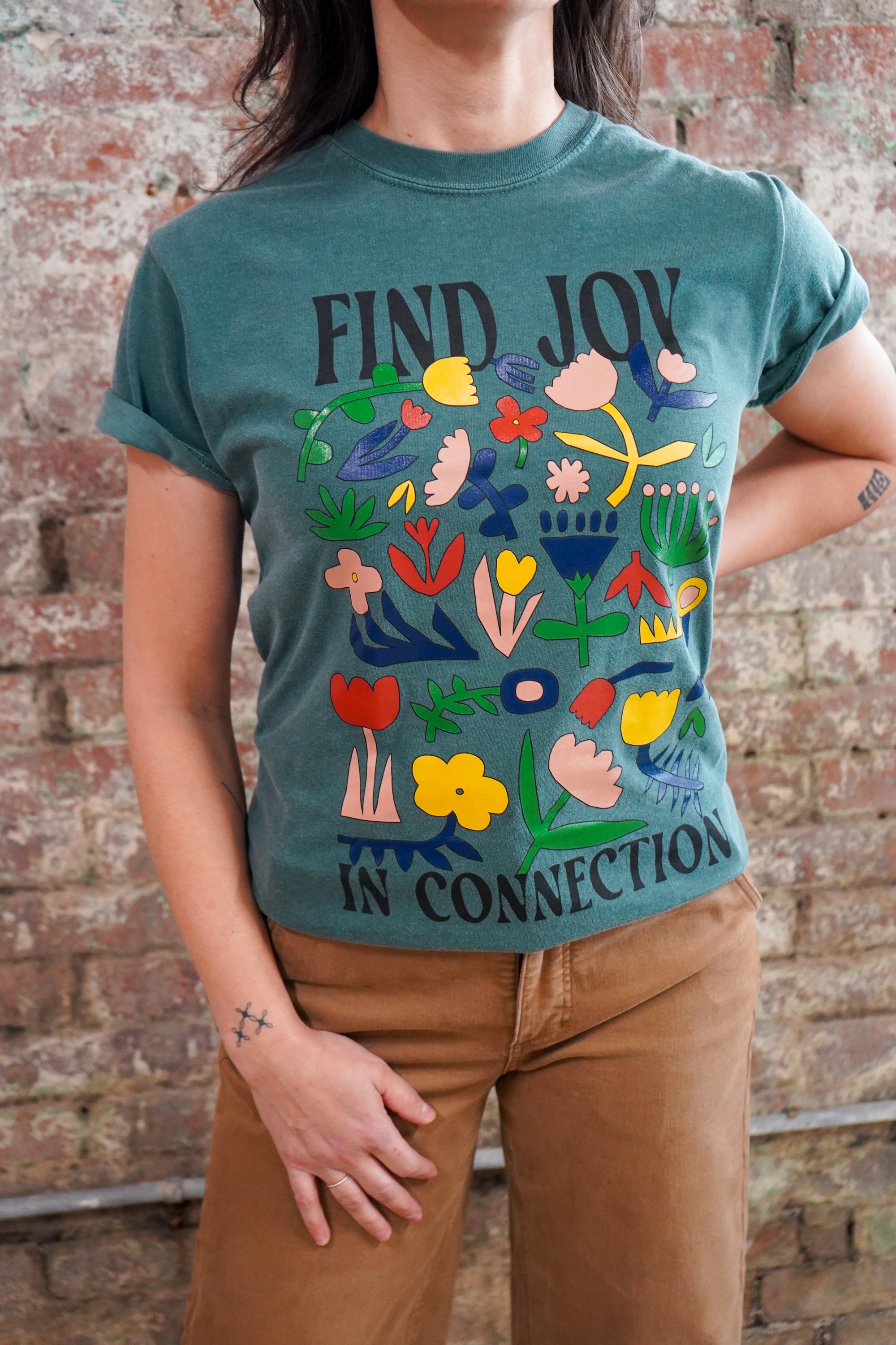 Person wearing a teal t-shirt with colorful designs and text against a brick wall.