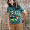 Woman wearing a green t-shirt with colorful graphics and text, standing against a brick wall.