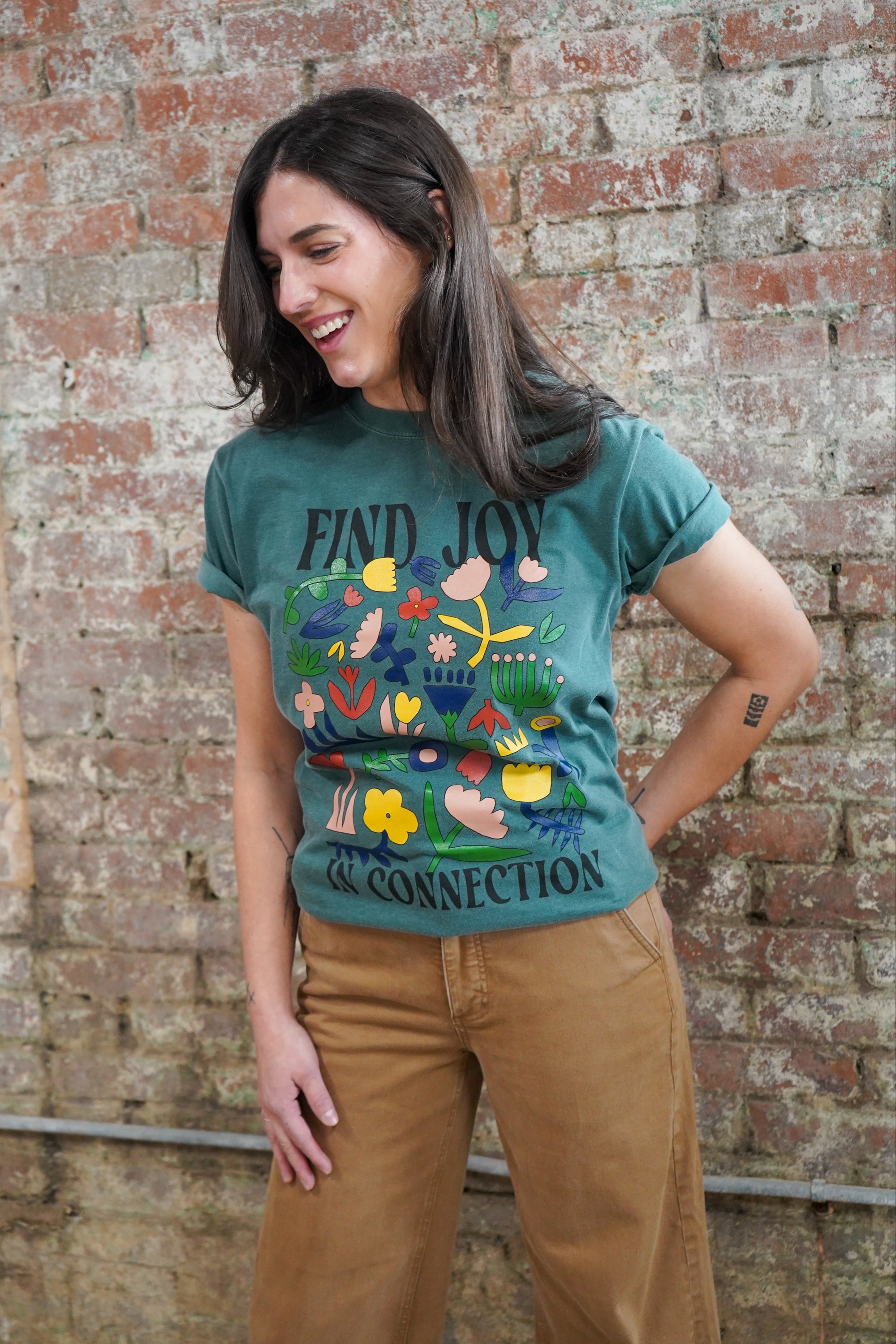 Woman wearing a green t-shirt with colorful graphics and text, standing against a brick wall.