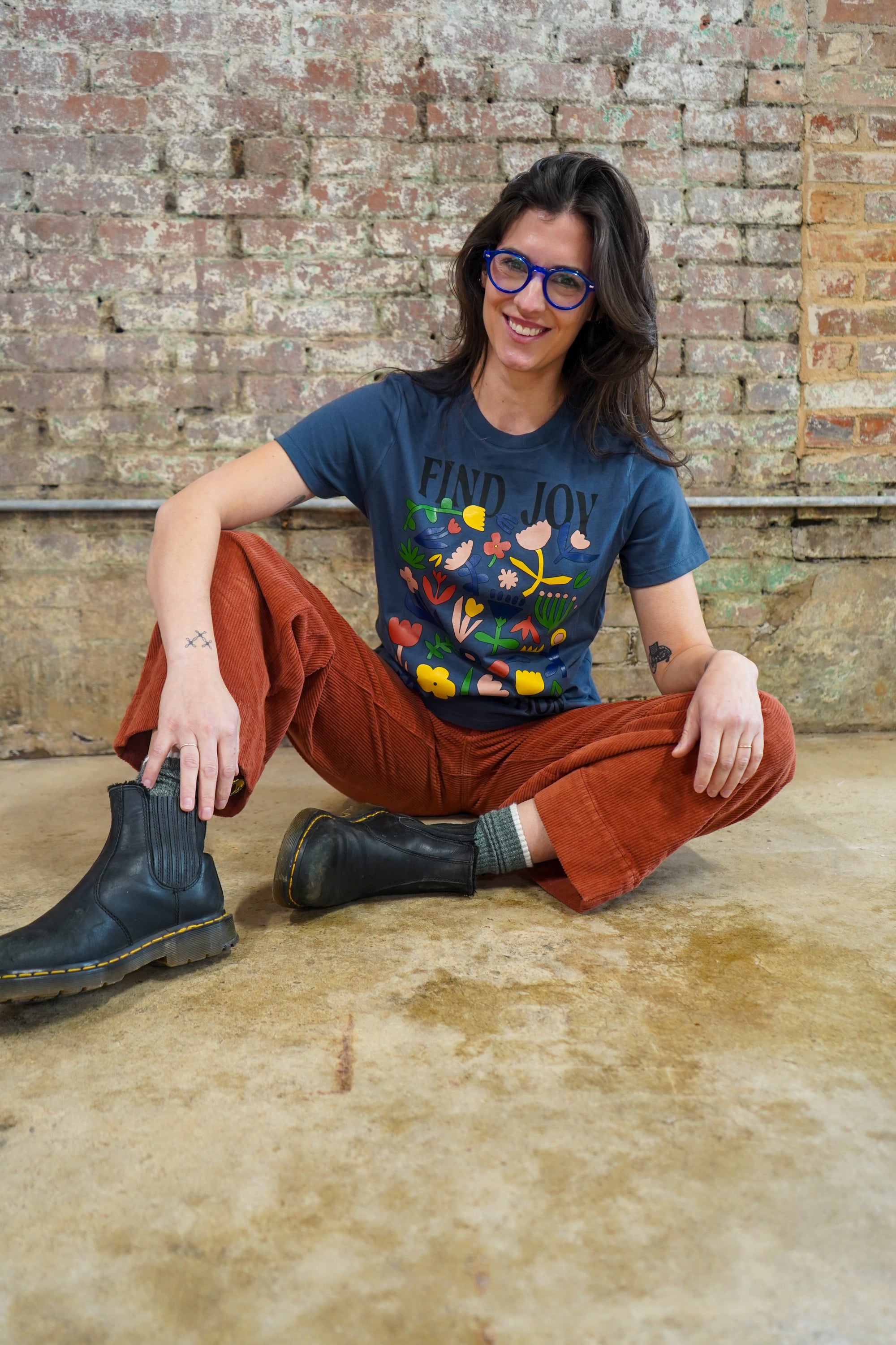 Person wearing a blue t-shirt with colorful design and red pants, sitting on a brick floor.
