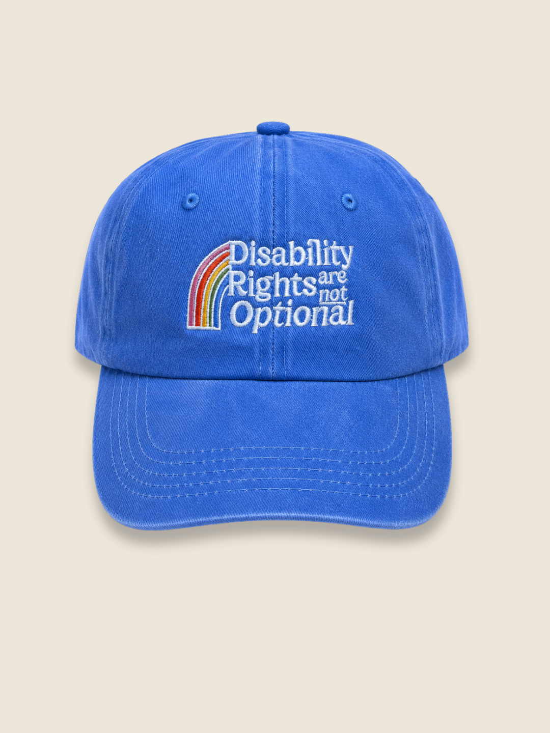 A blue baseball cap with an embroidered rainbow and the text 'Disability Rights are not Optional' on the front, set against a solid beige background.