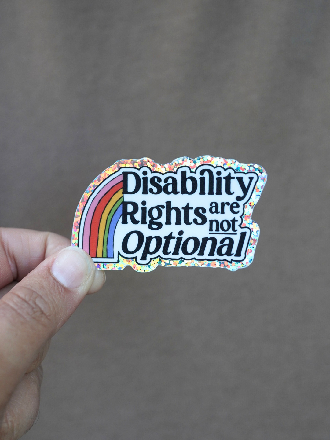 A hand holding a die-cut sticker that reads 'Disability Rights are not Optional' in black text over a rainbow graphic, with a holographic glitter border, against a plain brown background.