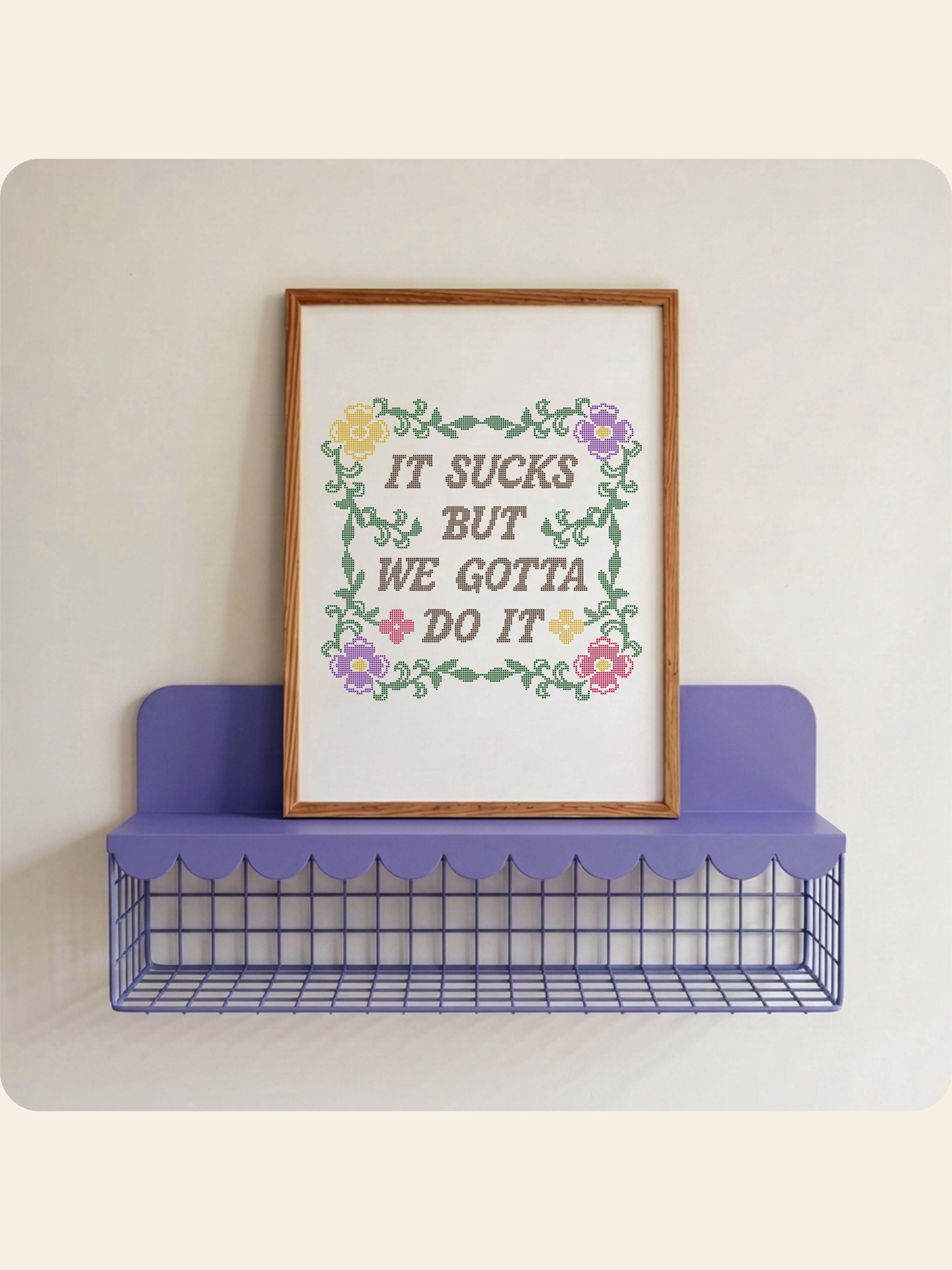 A framed motivational art print with the text 'IT SUCKS BUT WE GOTTA DO IT' surrounded by a colorful floral border, displayed on a purple wire shelf against a light beige wall.