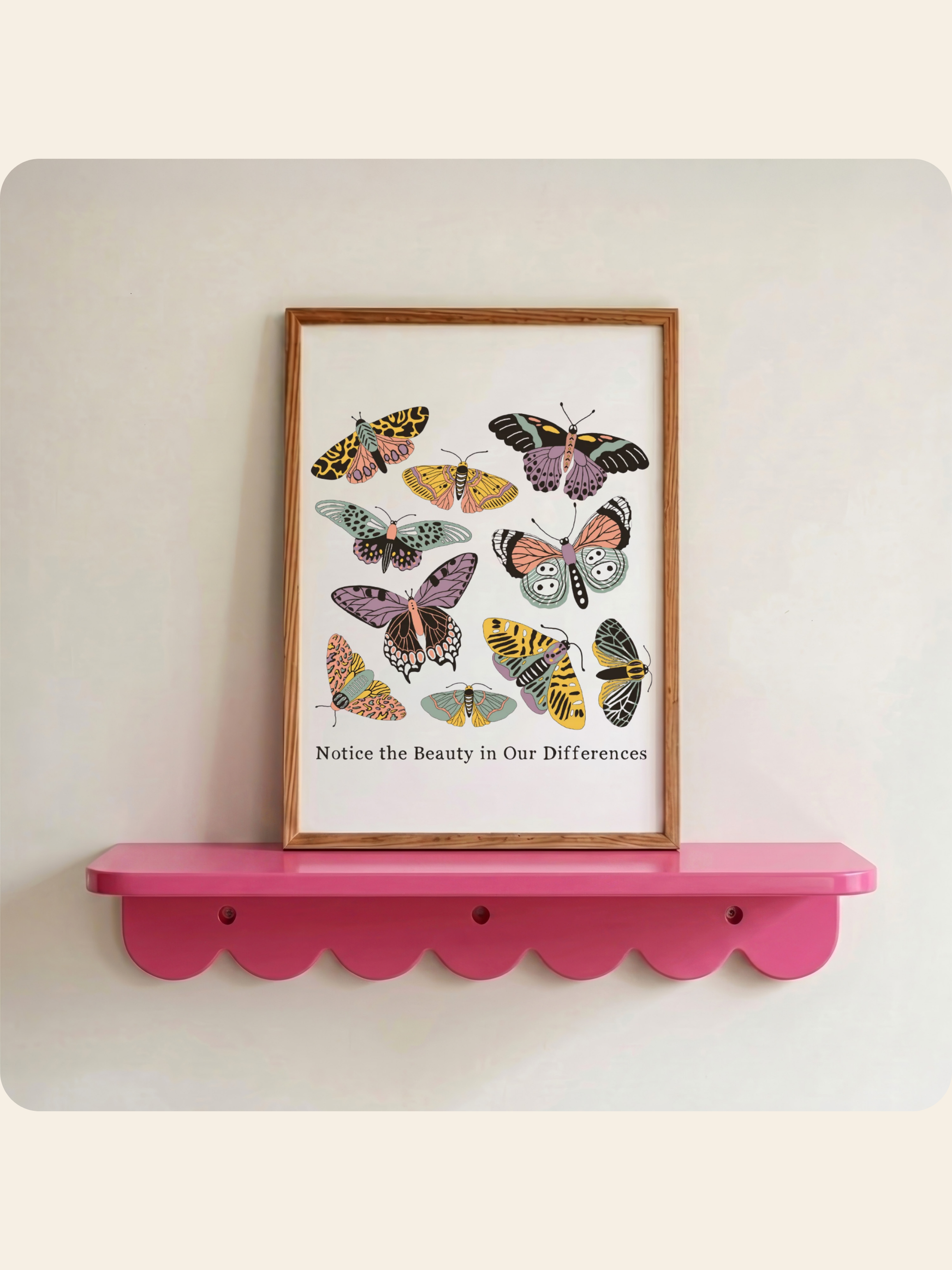 A framed butterfly art print with the text 'Notice the Beauty in Our Differences' displayed on a bright pink scalloped shelf against a light beige wall.