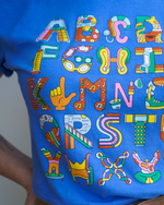 A close-up view of a blue t-shirt featuring a colorful graphic design of the alphabet. Each letter is uniquely decorated with various motifs and patterns, including musical notes, glasses, and other playful illustrations.