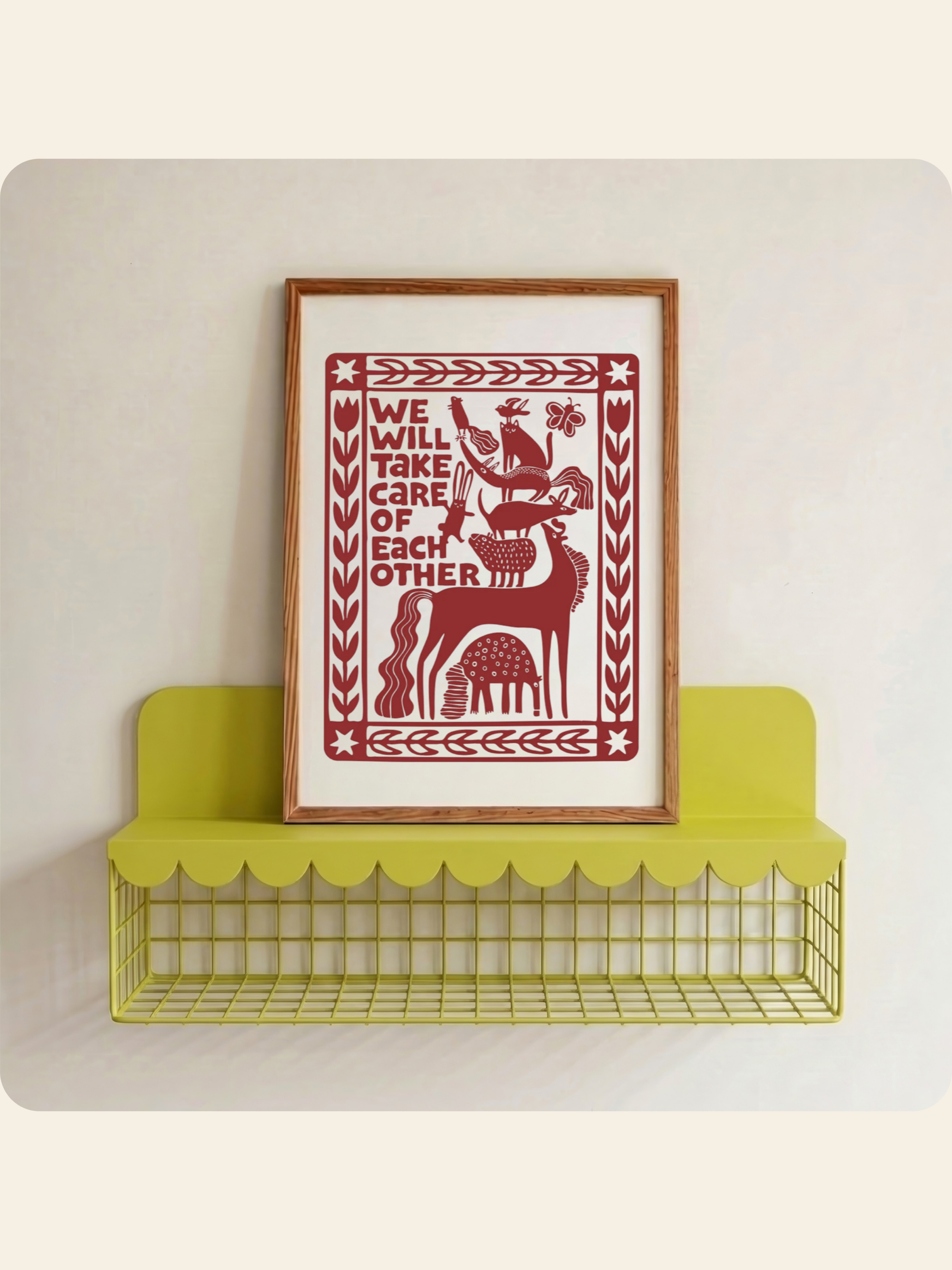 A framed art print with a red paper-cut style illustration of a deer and the text 'WE WILL TAKE CARE OF EACH OTHER' on a white background, leaning on a lime green wire shelf against a light beige wall.