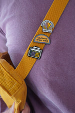 A close-up view of a yellow strap featuring three enamel pins. The pins read: 'MEET KIDS WHERE THEY ARE,' 'CELEBRATE SMALL WINS,' and an AAC device, all placed on a purple shirt.