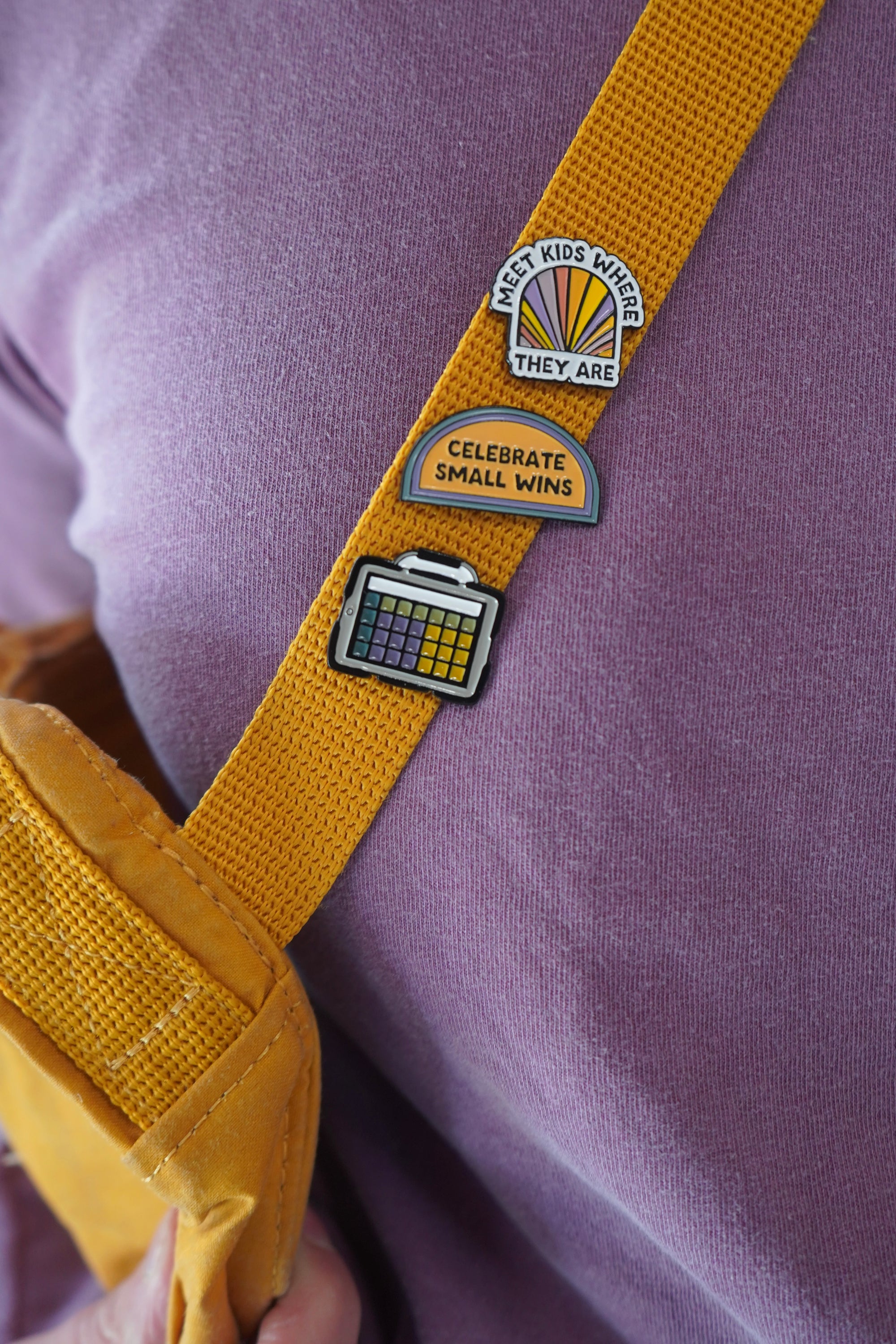 A close-up view of a yellow strap featuring three enamel pins. The pins read: 'MEET KIDS WHERE THEY ARE,' 'CELEBRATE SMALL WINS,' and an AAC device, all placed on a purple shirt.