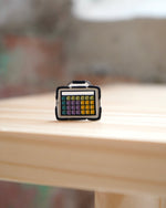 An enamel pin with a black base and multicolored segments, resembling aa AAC device, placed on a wooden surface.