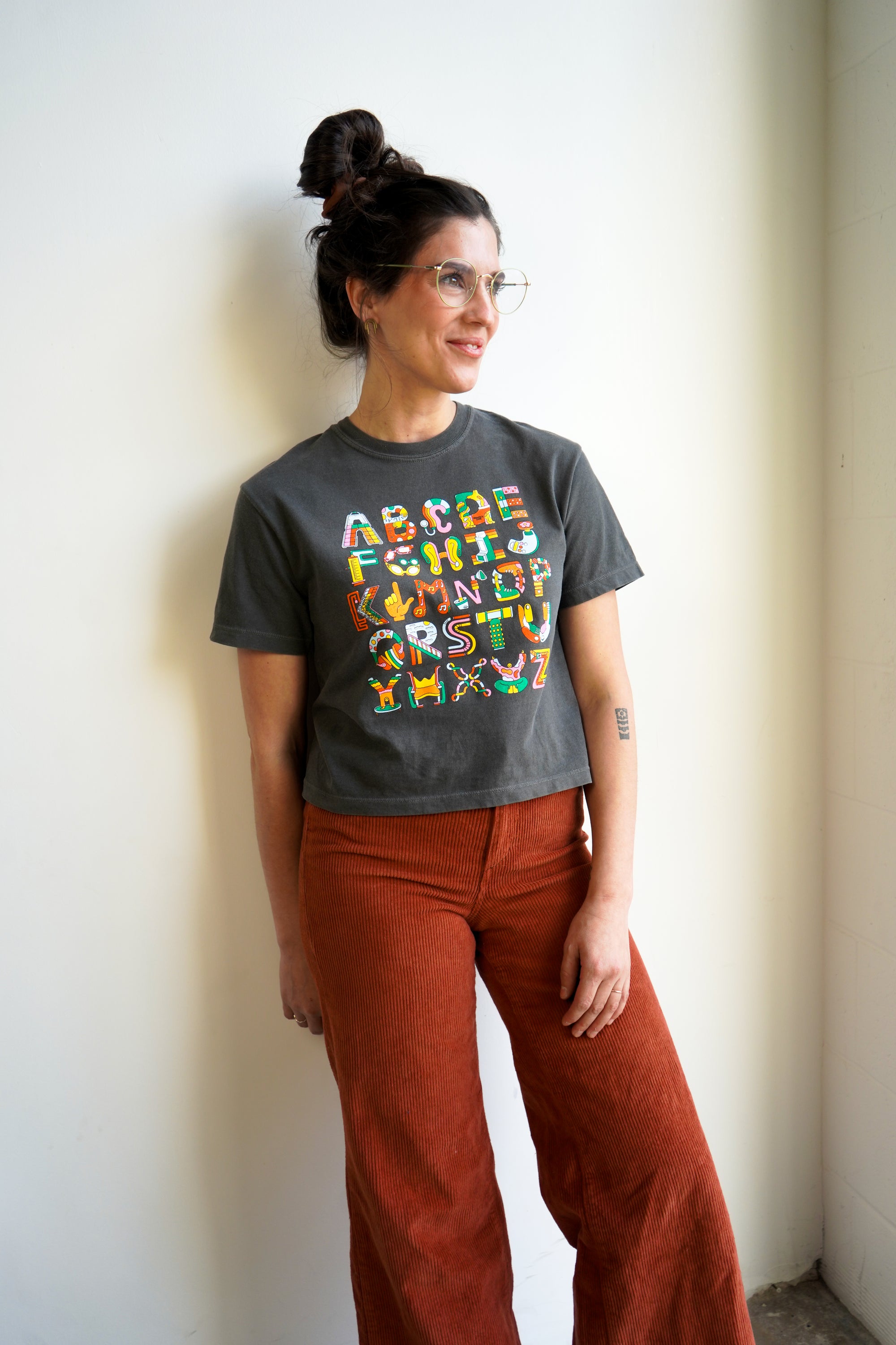 A woman with a bun hairstyle and glasses stands against a light wall. She wears a cropped gray t-shirt featuring colorful text and designs. Paired with rust-colored wide-leg pants, she has a tattoo on her arm and a relaxed posture.