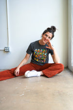 A woman with a bun hairstyle and glasses stands against a light wall. She wears a cropped gray t-shirt featuring colorful text and designs. Paired with rust-colored wide-leg pants, she has a tattoo on her arm and a relaxed posture.