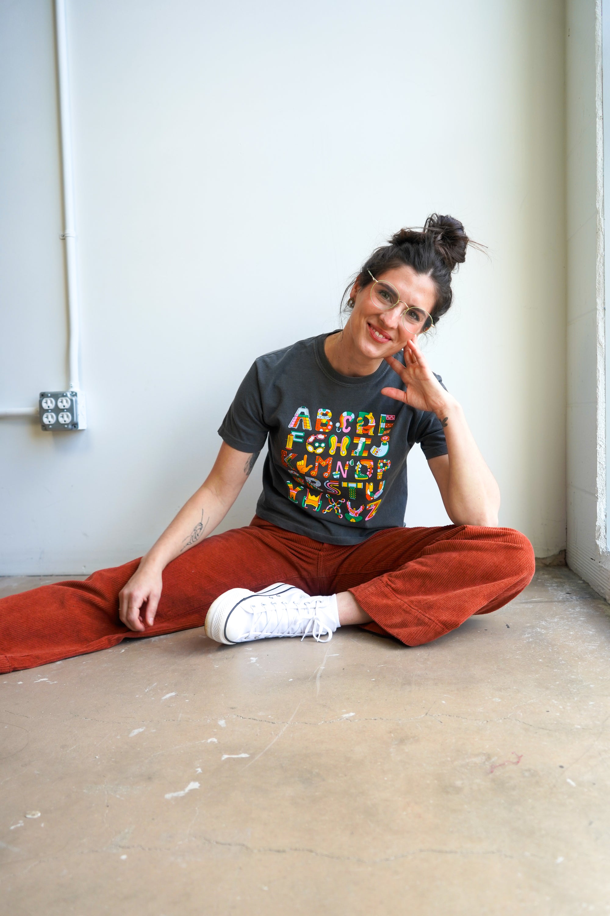 A woman with a bun hairstyle and glasses stands against a light wall. She wears a cropped gray t-shirt featuring colorful text and designs. Paired with rust-colored wide-leg pants, she has a tattoo on her arm and a relaxed posture.