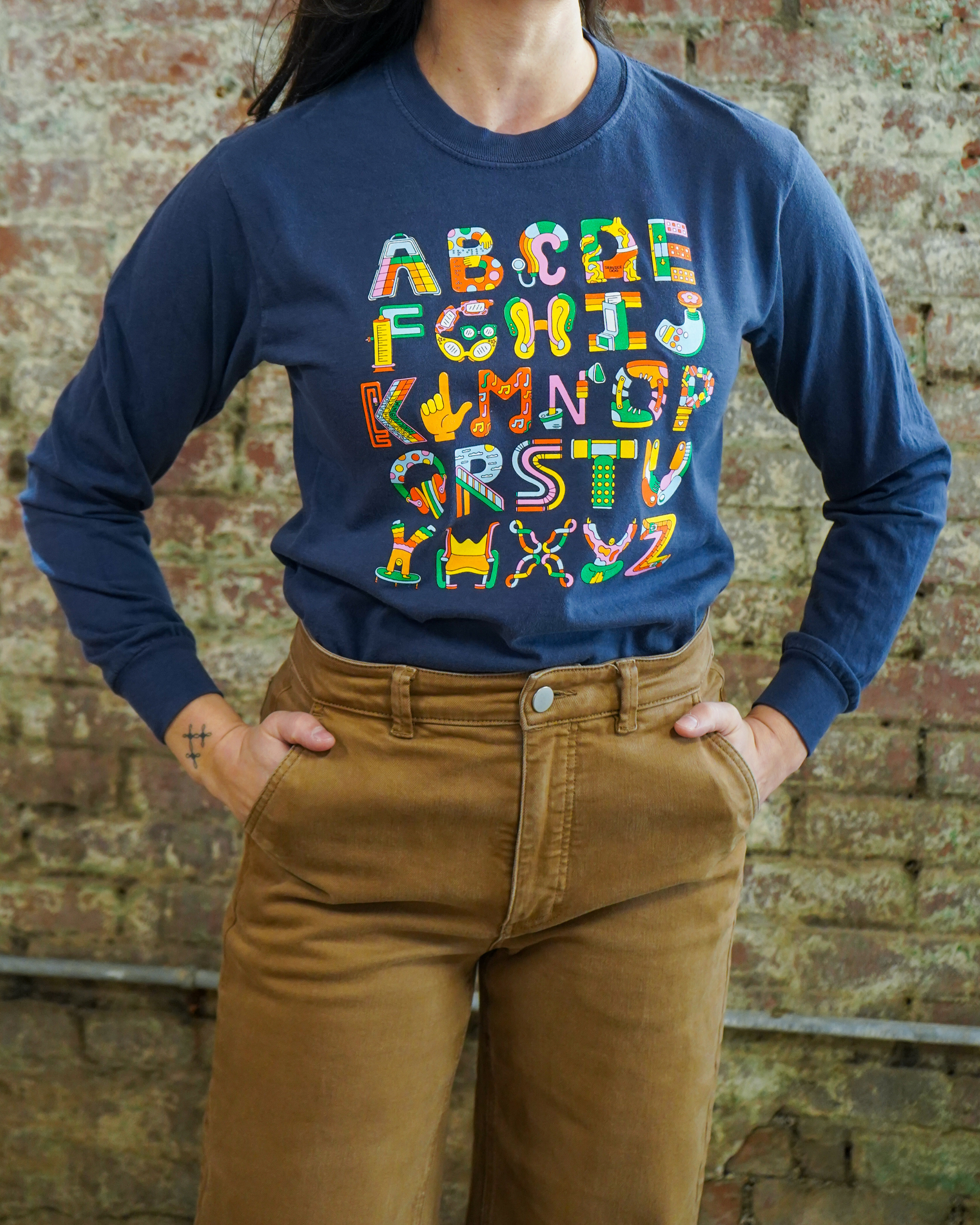 Person wearing a navy t shirt with colorful alphabet letters against a brick wall.