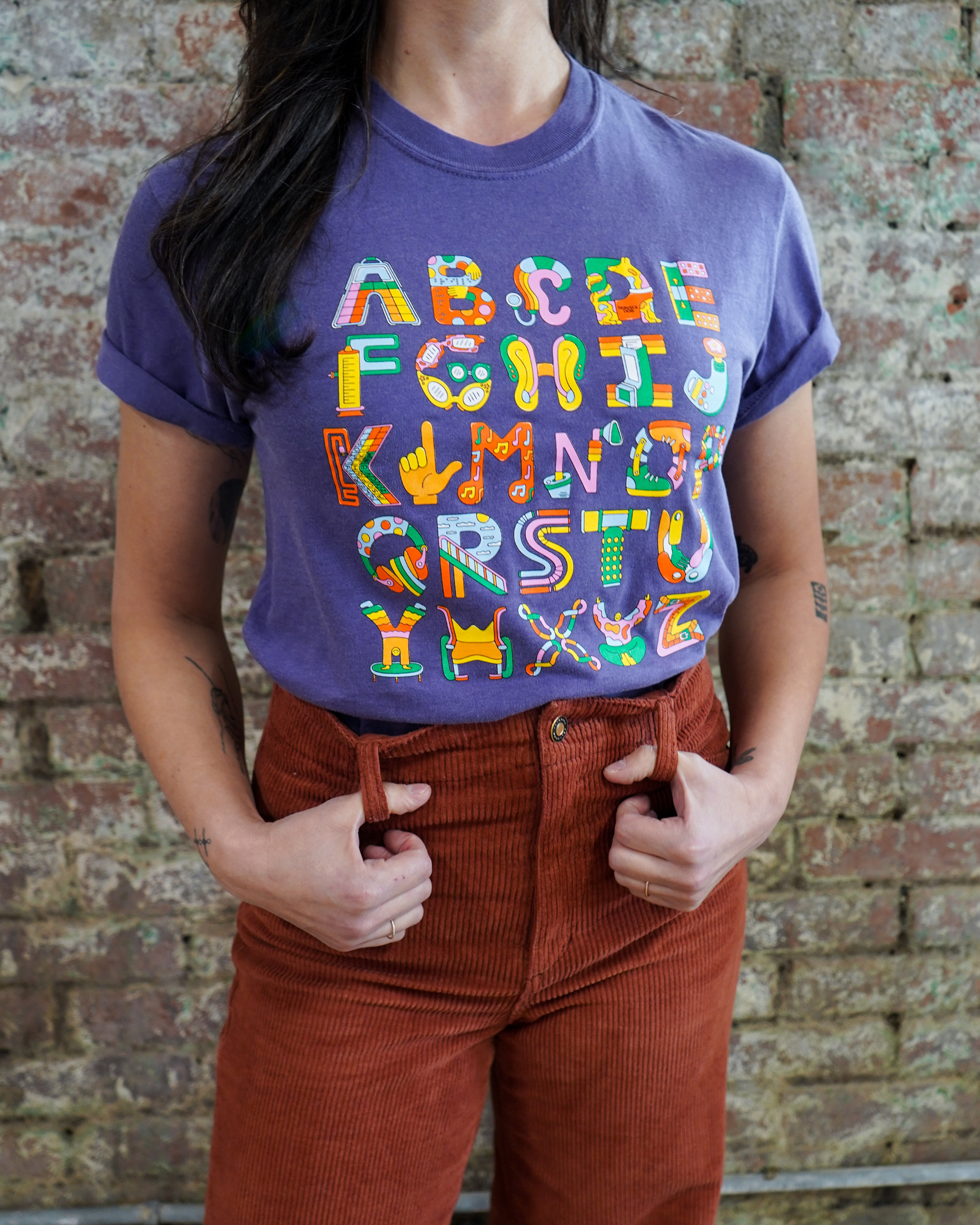 Person wearing a purple t-shirt with colorful letter design against a brick wall.