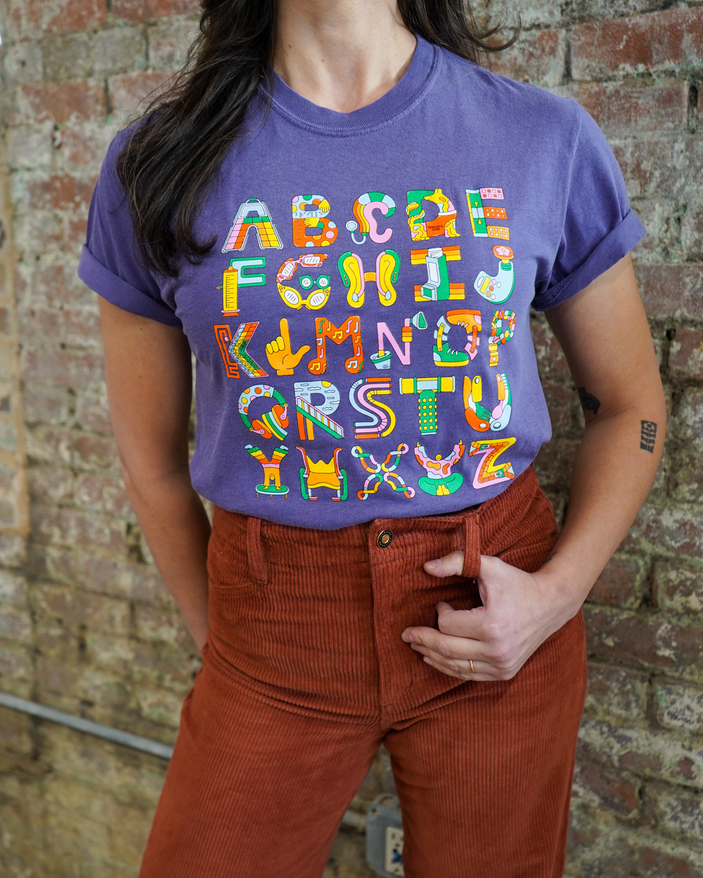Person wearing a purple t-shirt with colorful alphabet letters against a brick wall.