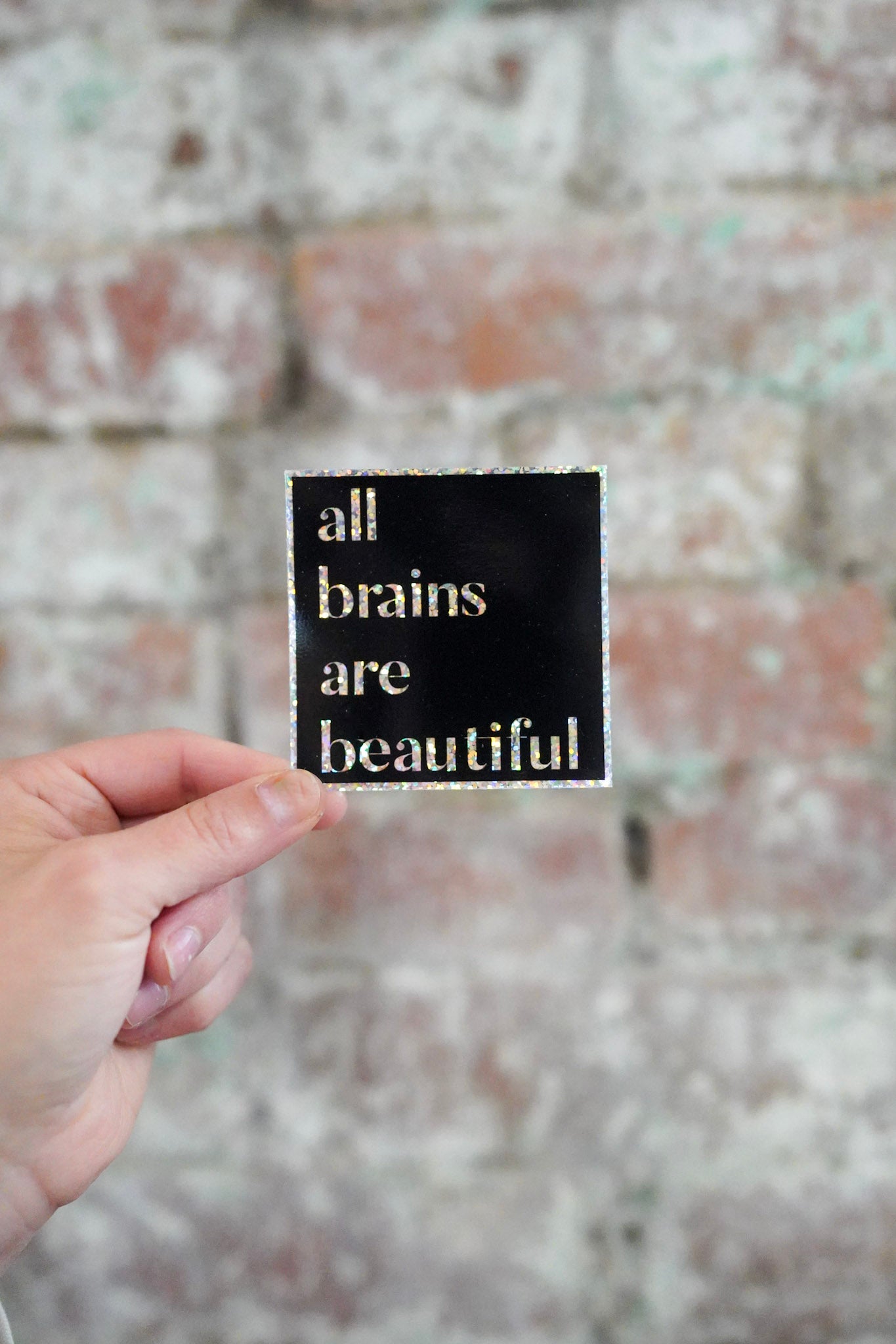 Hand holding a sign with 'all brains are beautiful' text against a brick wall.