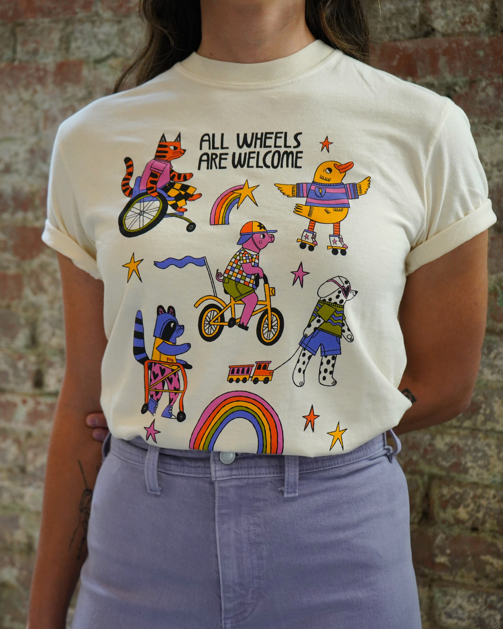 Person wearing a t-shirt with colorful illustrations and text against a brick wall.