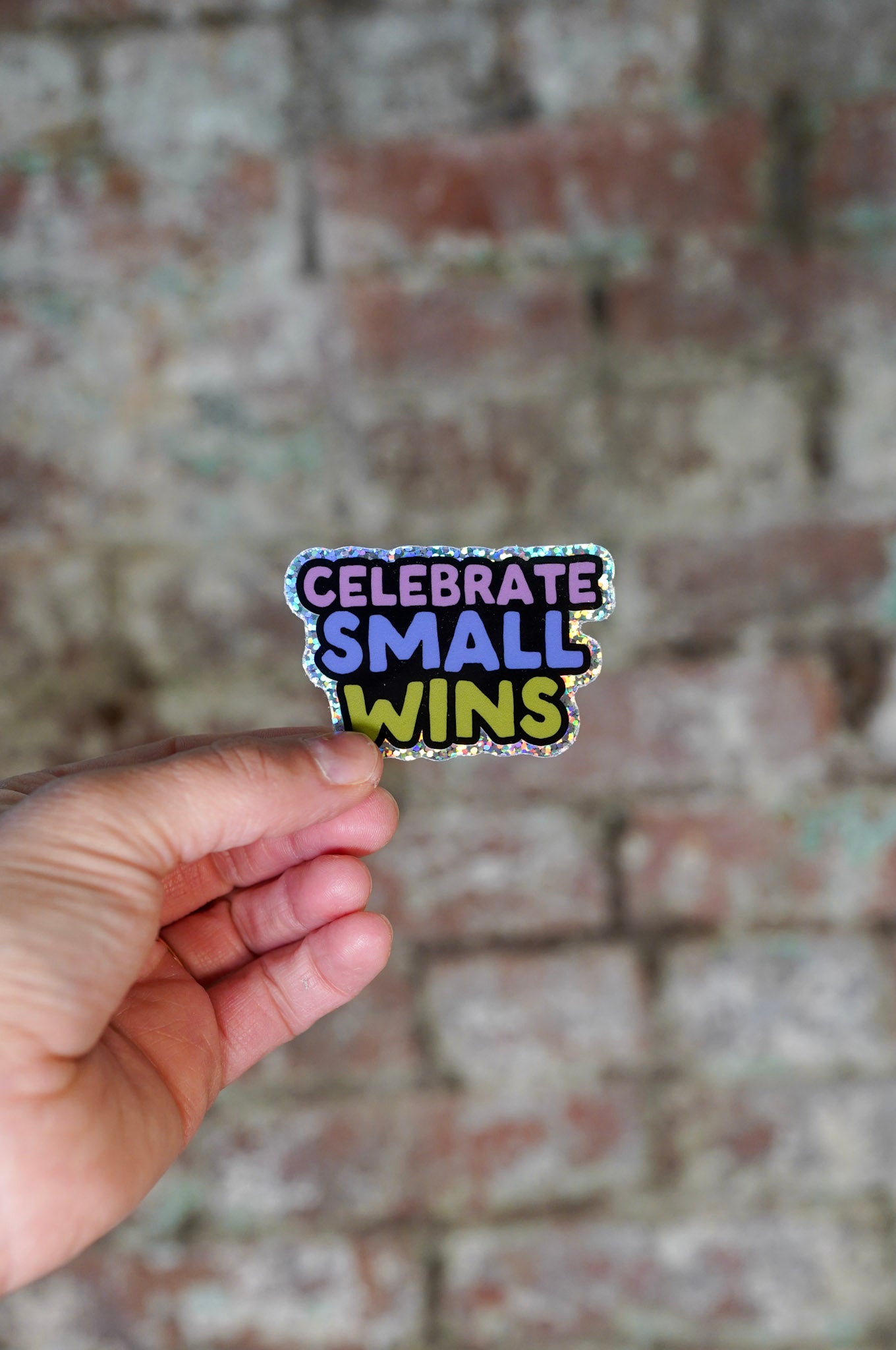 Hand holding a colorful sticker with 'Celebrate Small Wins' text against a brick wall background