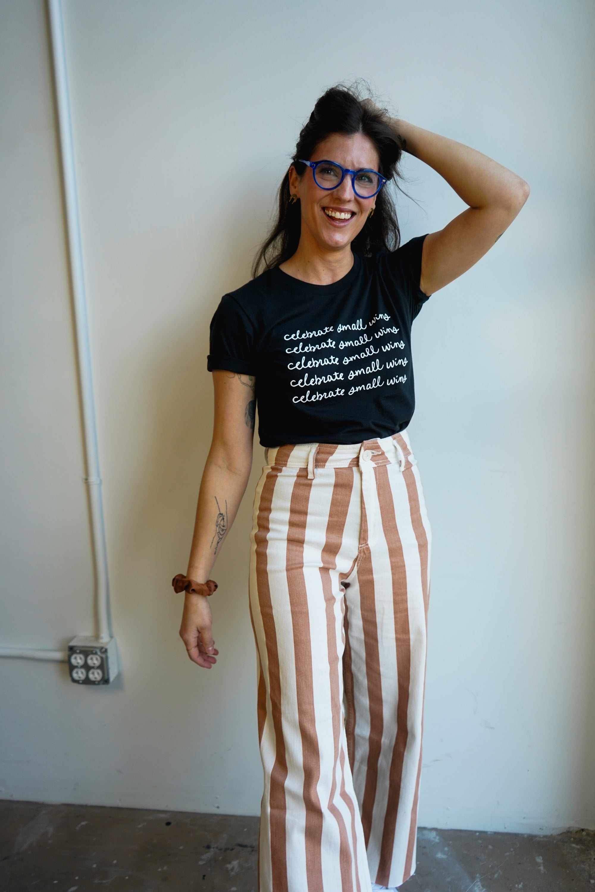 A woman wearing a black t-shirt with the phrase "celebrate small wins" printed in white repeatedly. She pairs it with high-waisted, striped brown and cream pants. The woman stands against a plain light-colored wall, showing a casual fashion style.