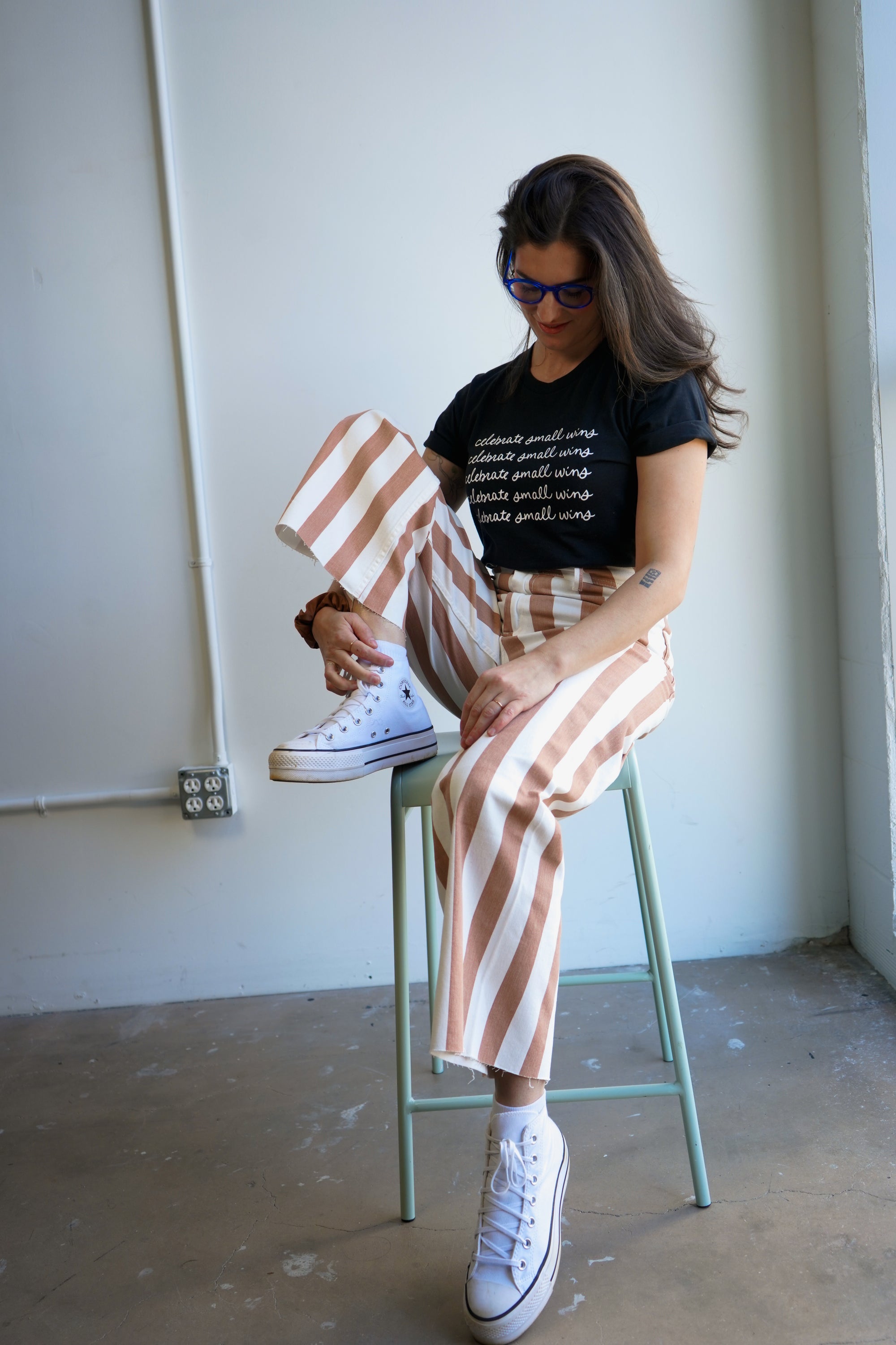 A woman wearing a black t-shirt with the phrase "celebrate small wins" printed in white repeatedly. She pairs it with high-waisted, striped brown and cream pants. The woman stands against a plain light-colored wall, showing a casual fashion style.