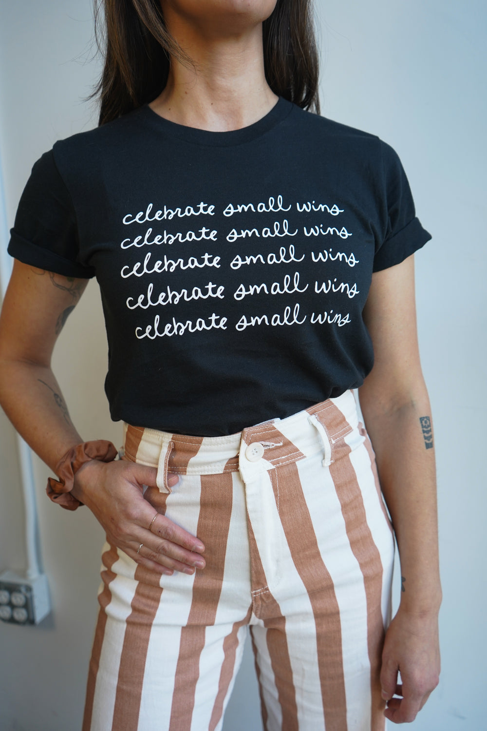 A woman wearing a black t-shirt with the phrase "celebrate small wins" printed in white repeatedly. She pairs it with high-waisted, striped brown and cream pants. The woman stands against a plain light-colored wall, showing a casual fashion style.