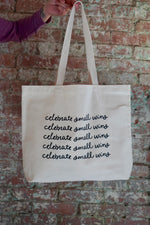 A person holding a large canvas tote bag with the phrase "celebrate small wins" printed multiple times in black cursive font. The background features a brick wall, and the person is wearing a purple long-sleeve shirt.