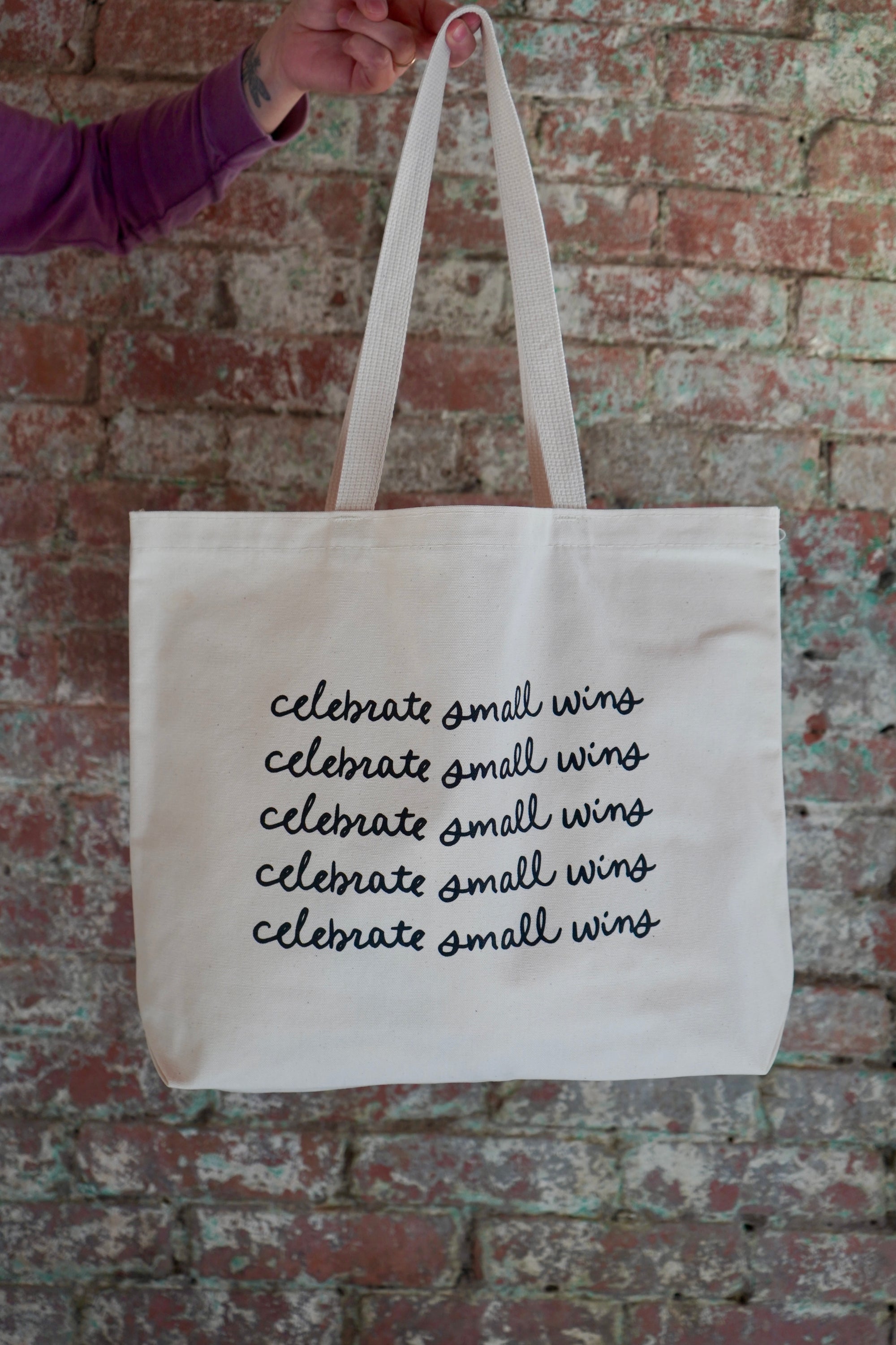 A person holding a large canvas tote bag with the phrase "celebrate small wins" printed multiple times in black cursive font. The background features a brick wall, and the person is wearing a purple long-sleeve shirt.