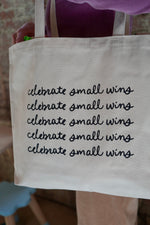 A person holding a large canvas tote bag with the phrase "celebrate small wins" printed multiple times in black cursive font. The background features a brick wall, and the person is wearing a purple long-sleeve shirt.