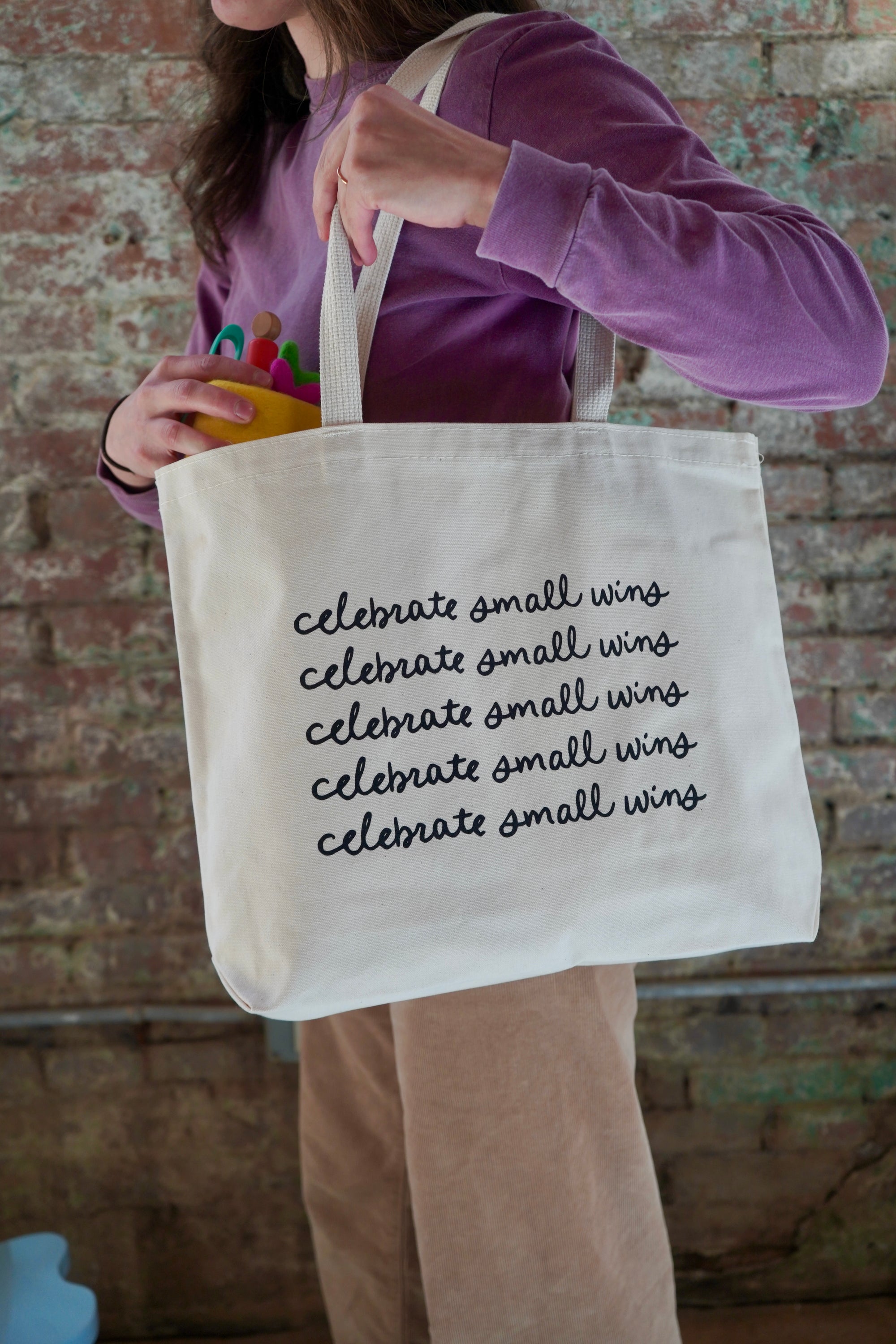 A person holding a large canvas tote bag with the phrase "celebrate small wins" printed multiple times in black cursive font. The background features a brick wall, and the person is wearing a purple long-sleeve shirt.