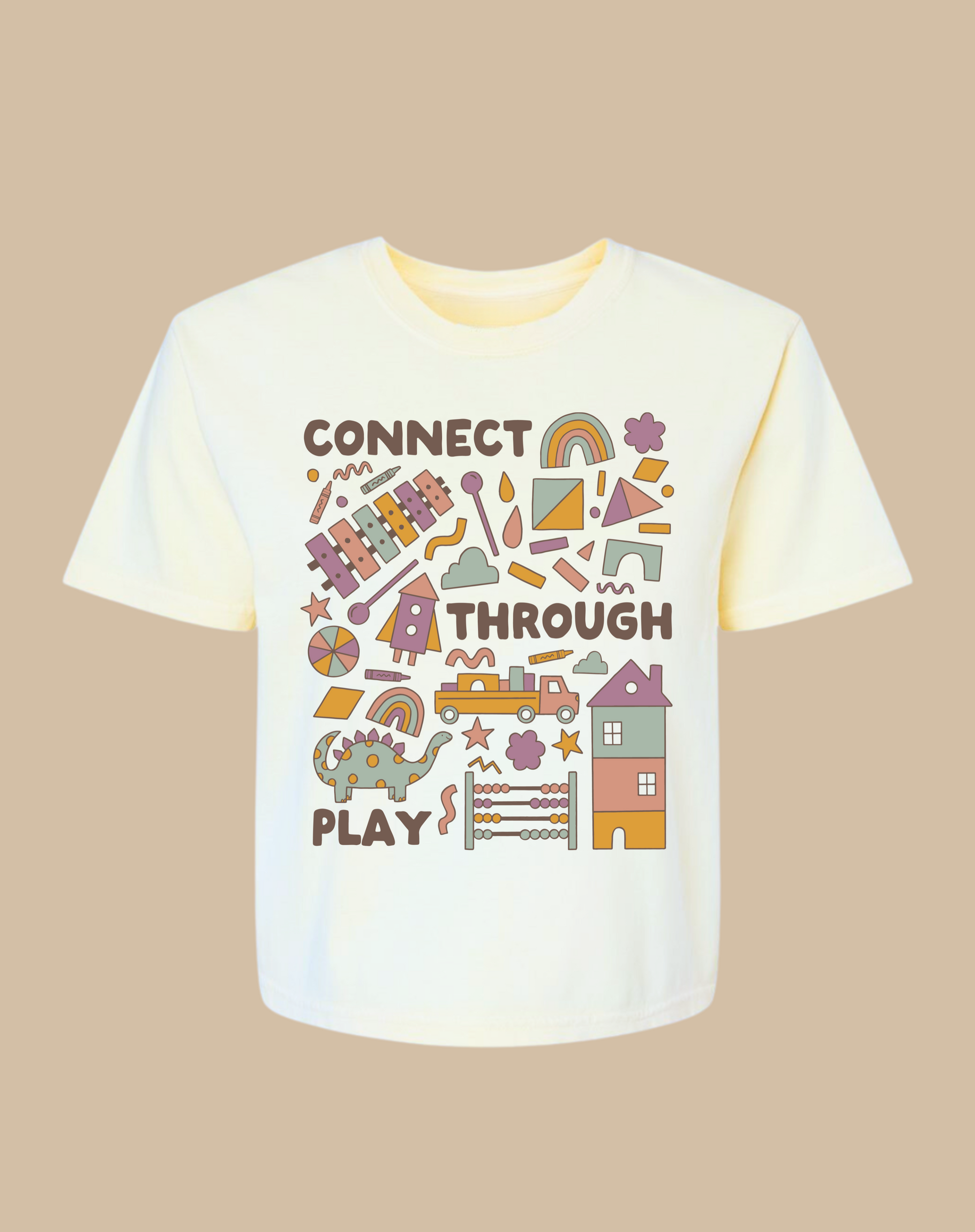 An ivory t-shirt featuring a playful graphic design with the words "CONNECT THROUGH PLAY." The design includes colorful shapes, a dinosaur, a rainbow, and various playful elements, set against a neutral background.