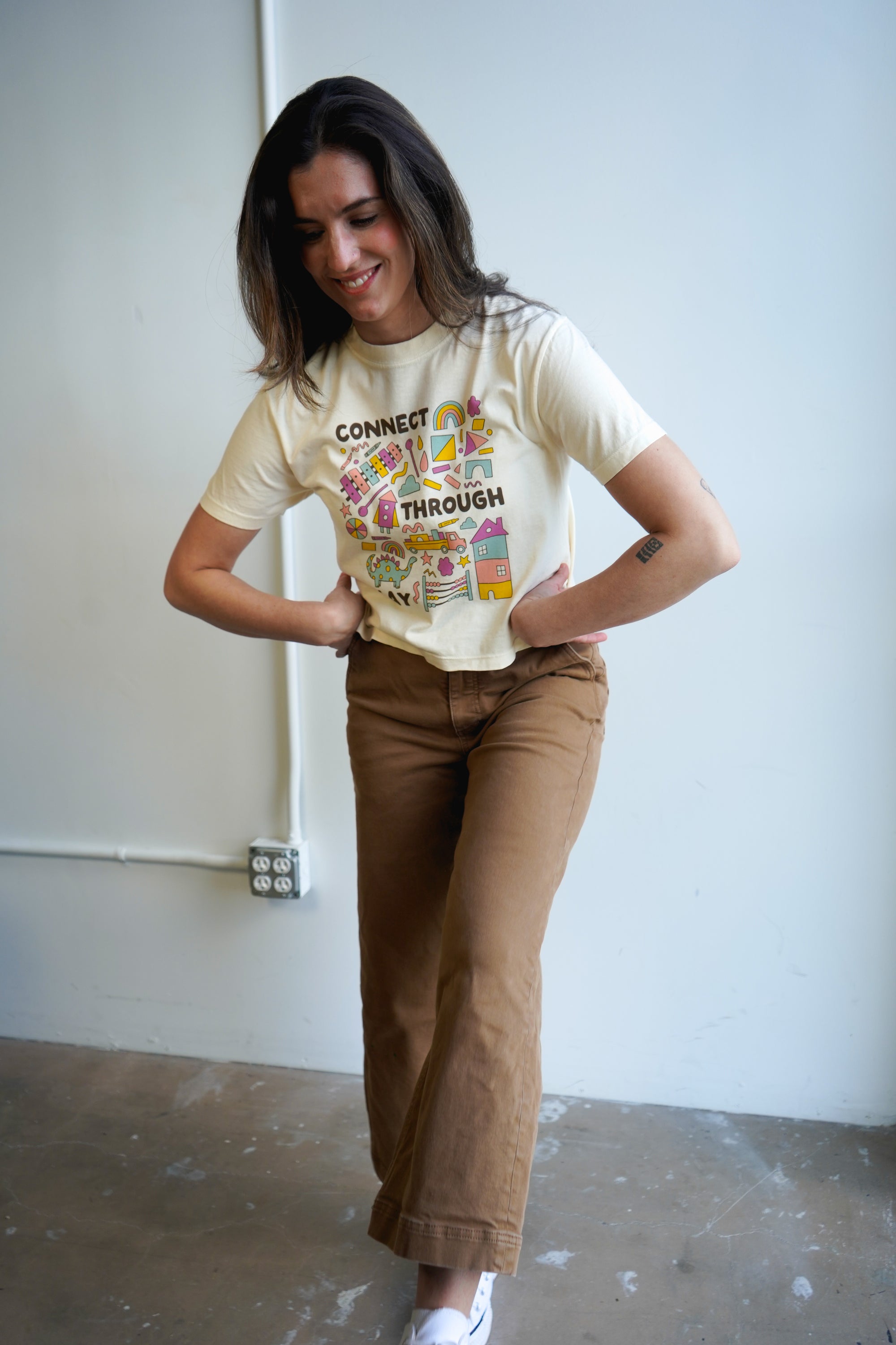 A woman wearing a light-colored t-shirt with the text "CONNECT THROUGH PLAY" surrounded by colorful illustrations of houses, rainbows, and playful elements. She is standing against a simple backdrop, wearing brown pants.