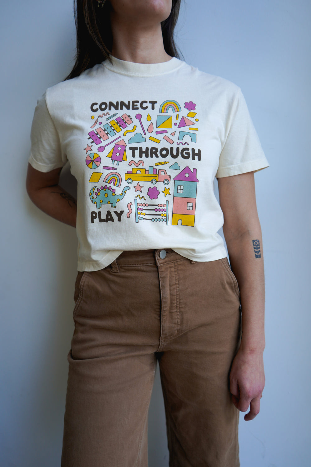 A woman wearing a light-colored t-shirt with the text "CONNECT THROUGH PLAY" surrounded by colorful illustrations of houses, rainbows, and playful elements. She is standing against a simple backdrop, wearing brown pants.