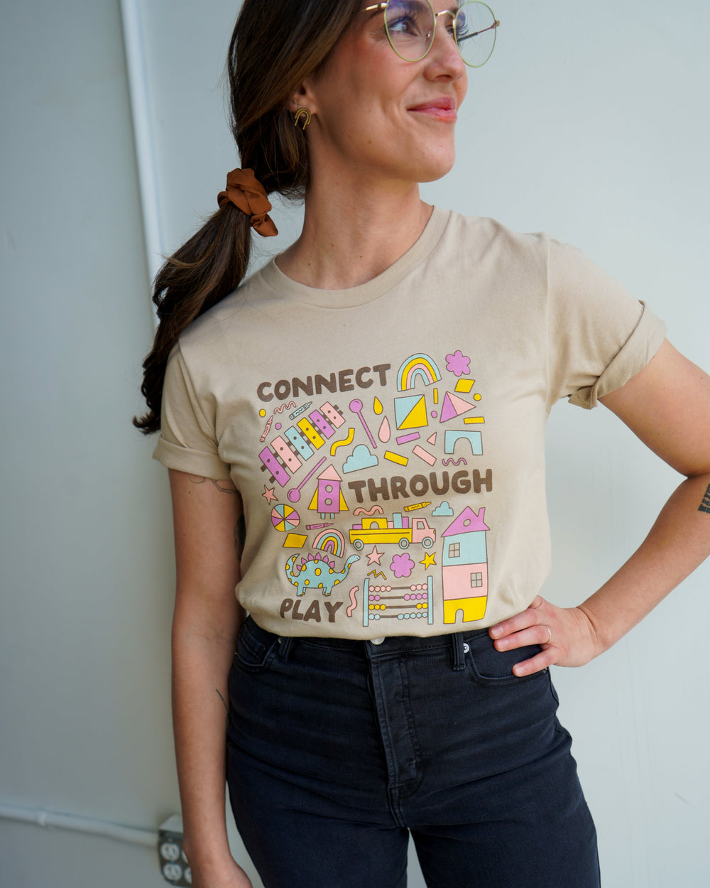 A person wearing a beige t-shirt with the text 'CONNECT THROUGH PLAY' and a colorful illustration of various playful icons.