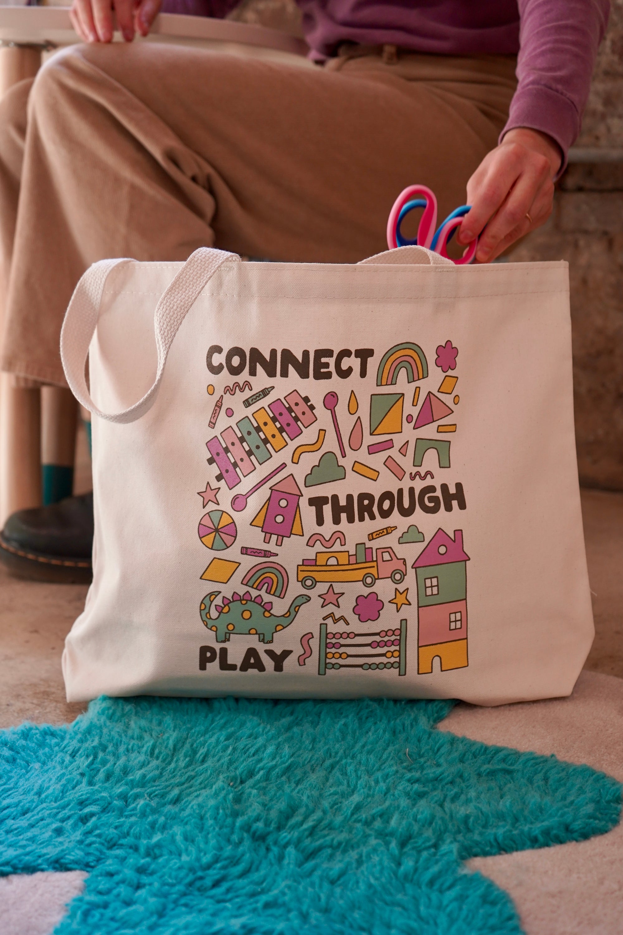 A cream-colored tote bag featuring colorful graphics with the text 'CONNECT THROUGH PLAY.' The bag is held by a person wearing a purple sweater, with scissors visible in their other hand. The background includes a turquoise rug and a wooden setting.