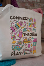 A cream-colored tote bag featuring colorful graphics with the text 'CONNECT THROUGH PLAY.' The bag is held by a person wearing a purple sweater, with scissors visible in their other hand. The background includes a turquoise rug and a wooden setting.