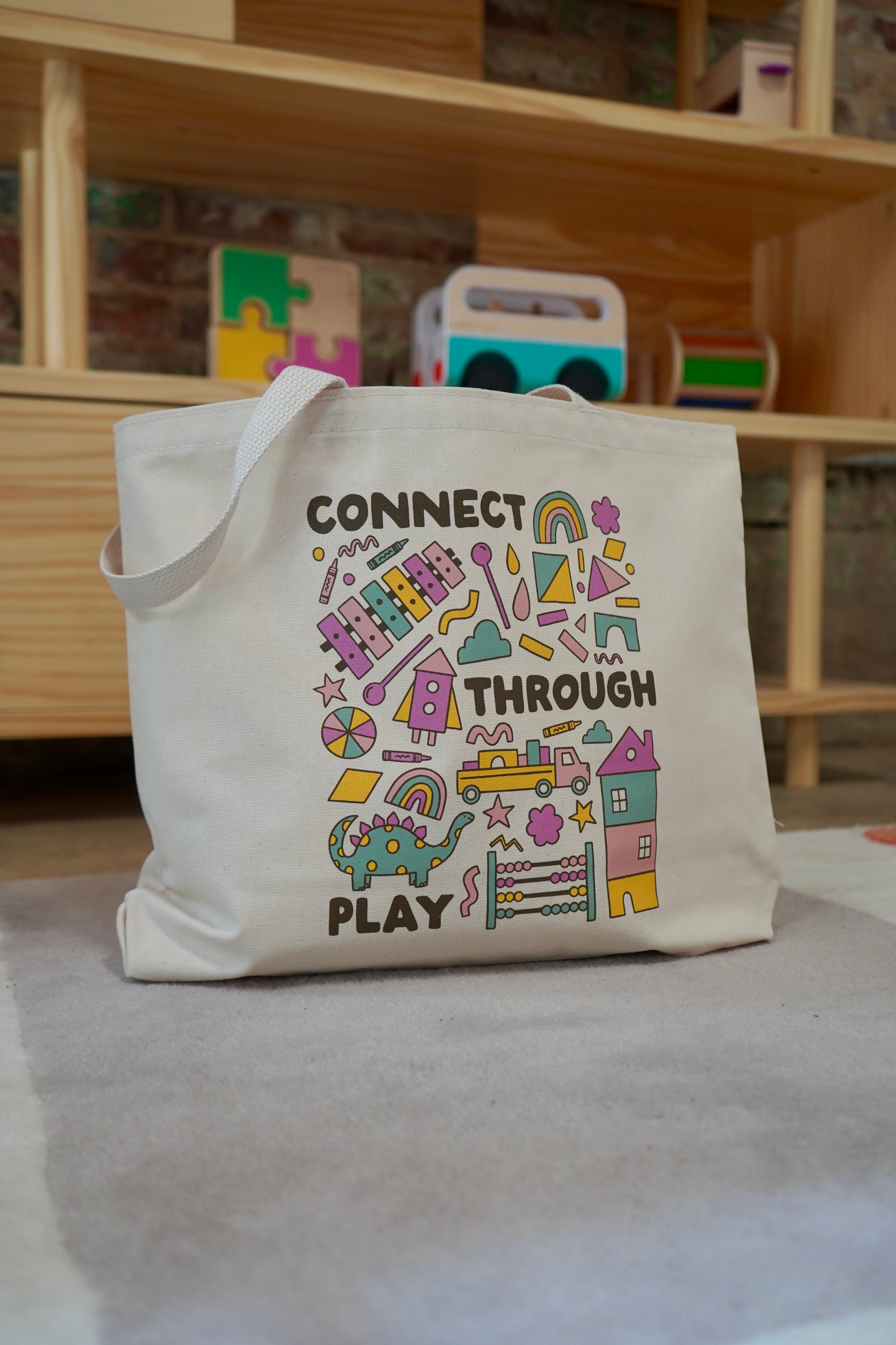 A canvas tote bag featuring colorful playful designs and the text "CONNECT THROUGH PLAY" in bold letters. The bag is placed on a light-colored carpet, with wooden shelves and colorful toys visible in the background.