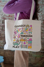 A person wearing a purple sweater is holding a white tote bag featuring colorful graphics and text that says "CONNECT THROUGH PLAY." The background consists of a textured brick wall.